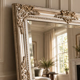 An ornate gold-framed floor mirror leaning against a beige paneled wall, reflecting a softly lit living room interior with curtains, a window, and plants.
