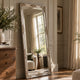 An ornate, full-length floor mirror with a carved silver frame leans against a beige paneled wall in a beautifully styled room with warm sunlight streaming through a window.