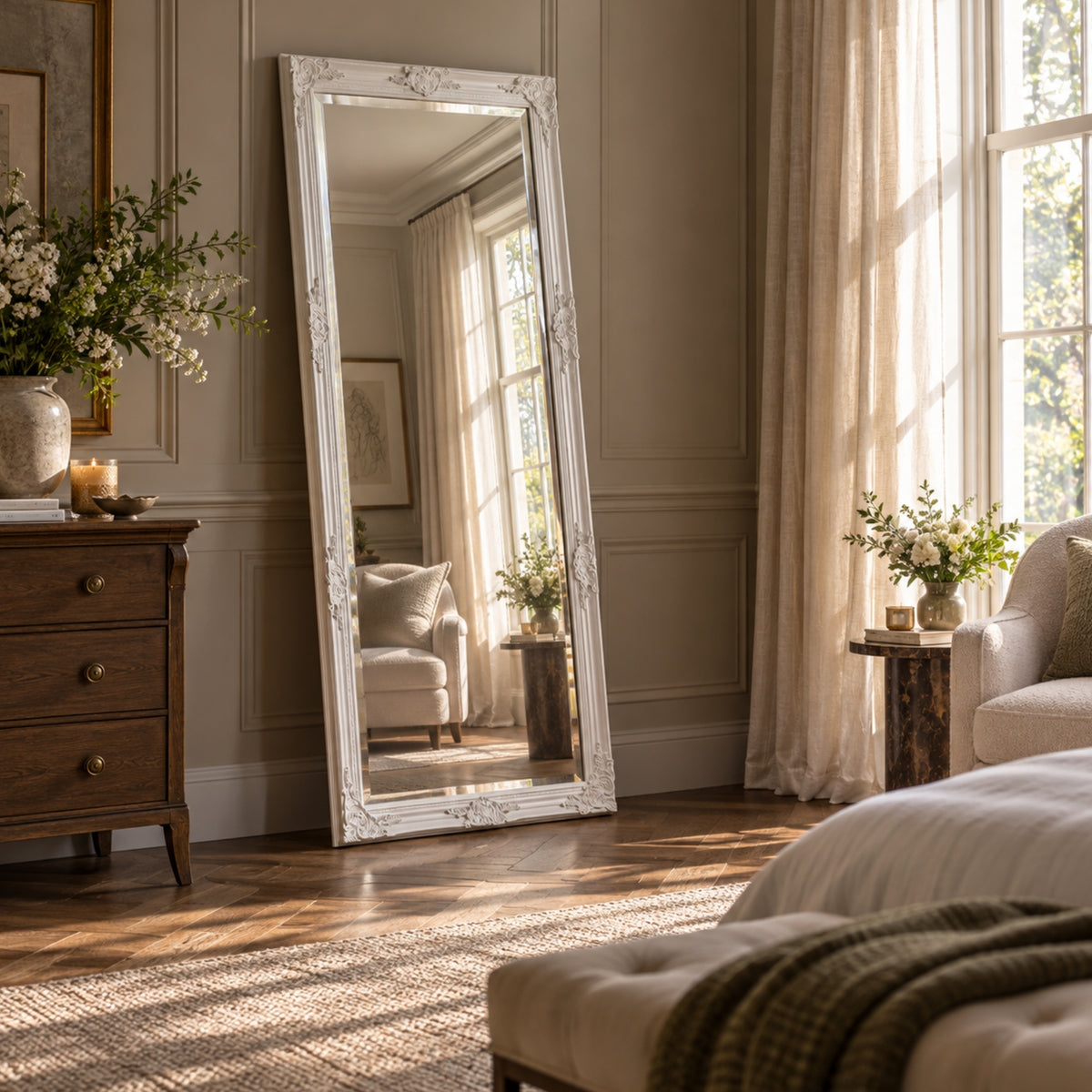 A tall floor mirror with an ornate white frame stands in a sunlit bedroom. The room has beige walls, wood floors, sheer curtains, a wooden chest, and a sofa, with the mirror reflecting part of the interior.