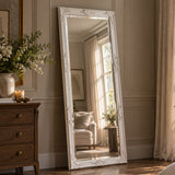 A tall floor mirror with an ornate white carved frame leans against a beige paneled wall in a sunlit room. The mirror reflects a softly lit chair and window, while a wooden dresser and floral arrangement are visible on the left.