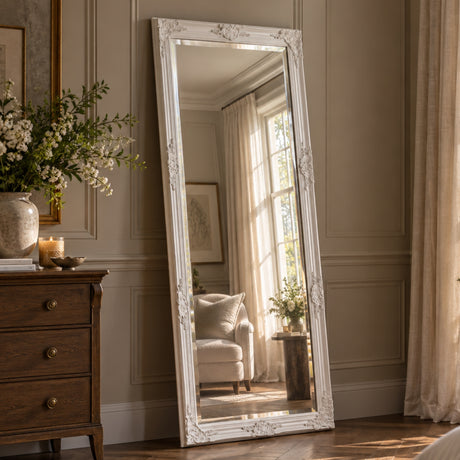 A tall floor mirror with an ornate white carved frame leans against a beige paneled wall in a sunlit room. The mirror reflects a softly lit chair and window, while a wooden dresser and floral arrangement are visible on the left.