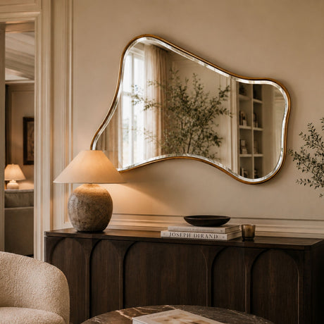 An organic-shaped wall mirror with a gold-toned frame hangs above a dark wood console table in a warm, styled interior. The mirror reflects a softly lit room with curtains and shelving, while the table below holds a lamp, books, and small decor items.