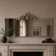An ornate rectangular wall mirror with a decorative silver-toned crest at the top, mounted above a fireplace mantel in a warm neutral room. The mirror reflects a window and framed artwork, while the mantel holds a dark vase with branches, a small bowl, and books.