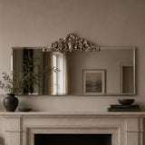 An ornate rectangular wall mirror with a decorative silver-toned crest at the top, mounted above a fireplace mantel in a warm neutral room. The mirror reflects a window and framed artwork, while the mantel holds a dark vase with branches, a small bowl, and books.