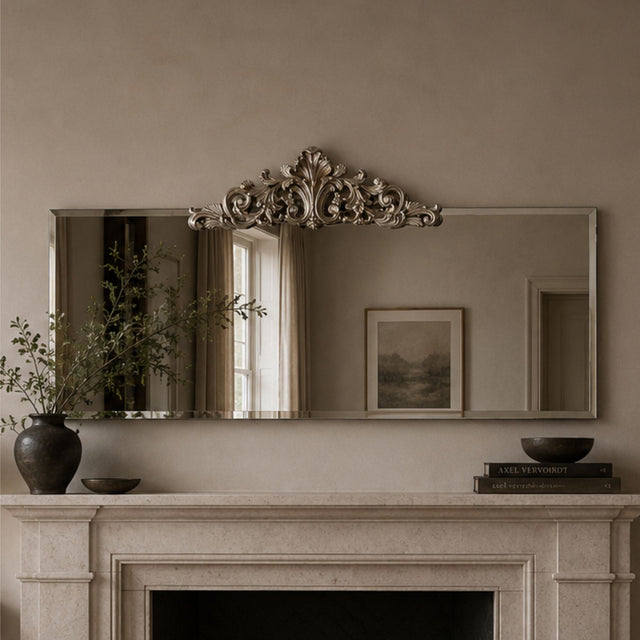 An ornate rectangular wall mirror with a decorative silver-toned crest at the top, mounted above a fireplace mantel in a warm neutral room. The mirror reflects a window and framed artwork, while the mantel holds a dark vase with branches, a small bowl, and books.