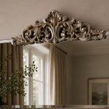 An ornate silver mirror frame with a highly detailed baroque-style acanthus leaf and scrollwork top, shown reflecting a neutral-toned room with beige curtains and a window.