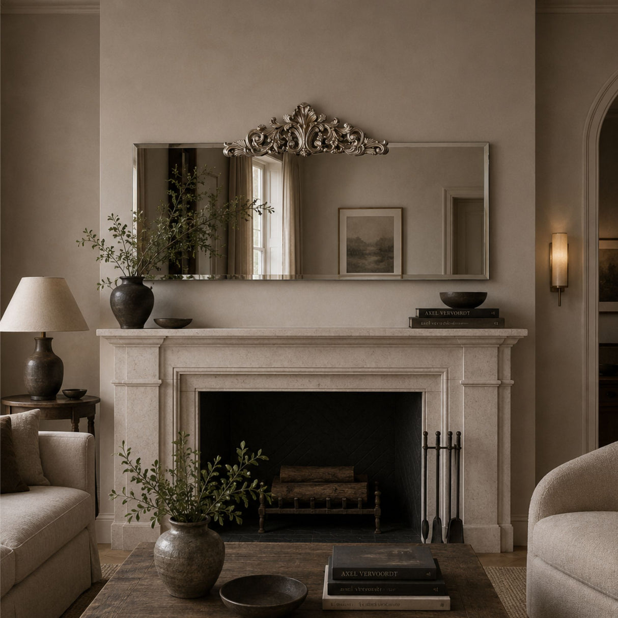 A styled interior scene featuring a large rectangular wall mirror with an ornate silver crest above a white stone fireplace mantel. The room has warm beige walls, a lit wall sconce, a side table with a lamp and vase, and upholstered seating partially visible at the sides.