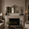 A styled interior scene featuring a large rectangular wall mirror with an ornate silver crest above a white stone fireplace mantel. The room has warm beige walls, a lit wall sconce, a side table with a lamp and vase, and upholstered seating partially visible at the sides.
