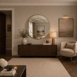 A styled interior room scene featuring a large round gold-framed mirror mounted on a beige wall above a dark wood console. The room includes a cream armchair, a textured rug, a side table with books and flowers, and framed wall art.