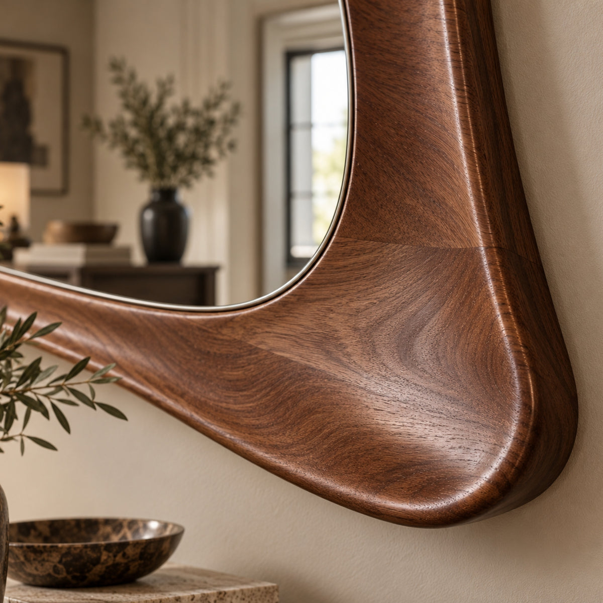 A close-up of a wall-mounted mirror with a thick, curved wooden frame in a warm medium-to-dark brown finish, set against a beige wall in a softly blurred interior room.