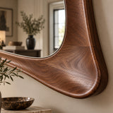 A close-up of a wall-mounted mirror with a thick, curved wooden frame in a warm medium-to-dark brown finish, set against a beige wall in a softly blurred interior room.