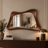 An organic-shaped wall mirror with a rich brown leather frame, mounted on a beige wall above a dark wood console table decorated with books, a vase, and a brass table lamp.