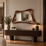 An organic-shaped brown leather wall mirror with a wavy, asymmetrical frame, mounted on a beige wall above a dark wood console table in a beautifully styled room.
