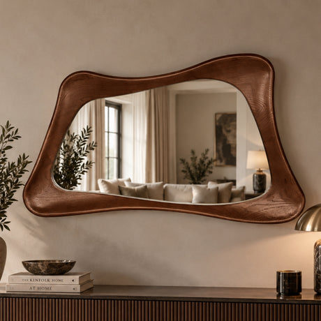 An organic-shaped wooden wall mirror with a rich brown textured frame, mounted on a beige wall above a console table in a styled living room setting.