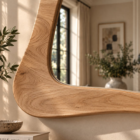 A large, uniquely shaped wooden wall mirror with a curved, organic frame, shown mounted on a beige wall in a beautifully lit room with soft sunlight and blurred interior decor.