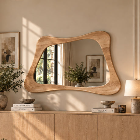 An organic-shaped wooden wall mirror with a wavy, hand-carved frame, mounted on a beige wall above a wooden credenza in a beautifully styled room.