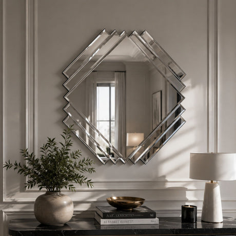 A large geometric diamond-shaped wall mirror with a silver mirrored frame hangs on a light gray paneled wall above a dark marble console table styled with a potted plant, books, a bowl, and a table lamp.