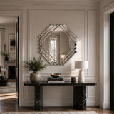 A large geometric octagonal mirror with a silver mirrored frame hangs on a beige paneled wall above a black marble console table in a styled entryway.