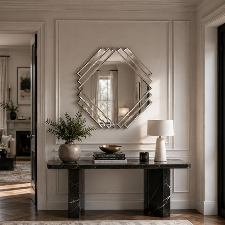 A large geometric octagonal mirror with a silver mirrored frame hangs on a beige paneled wall above a black marble console table in a styled entryway.