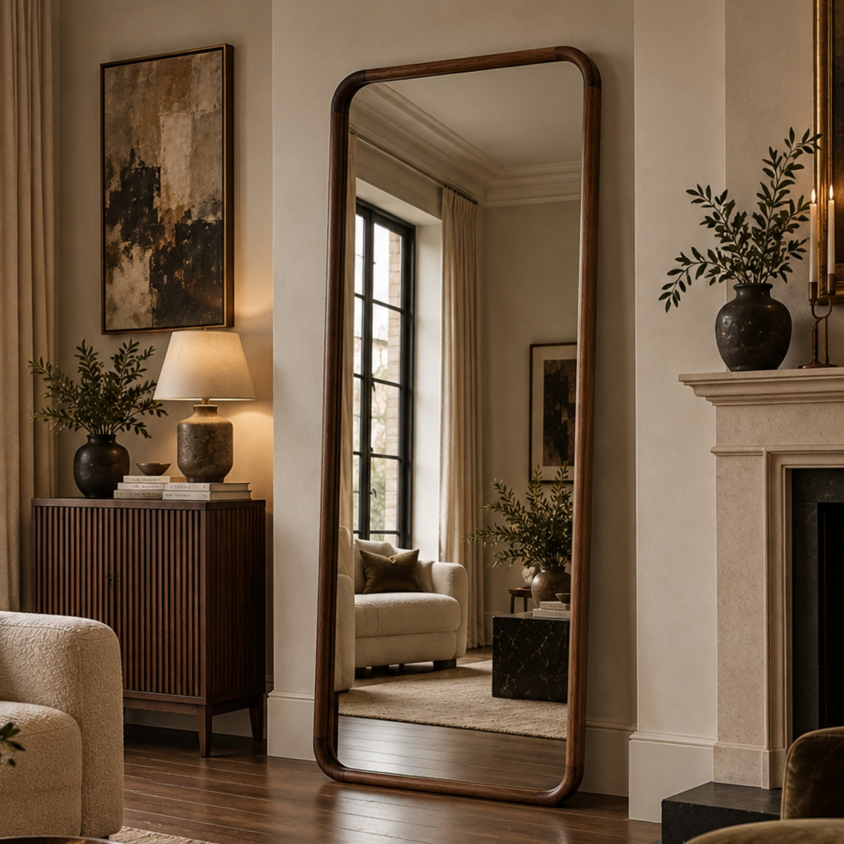 A tall floor mirror with a dark wood frame leans against a wall in a styled living room, reflecting a window, sofa, and artwork. The room features beige walls, a wooden cabinet, a lamp, and a fireplace mantel with potted plants.