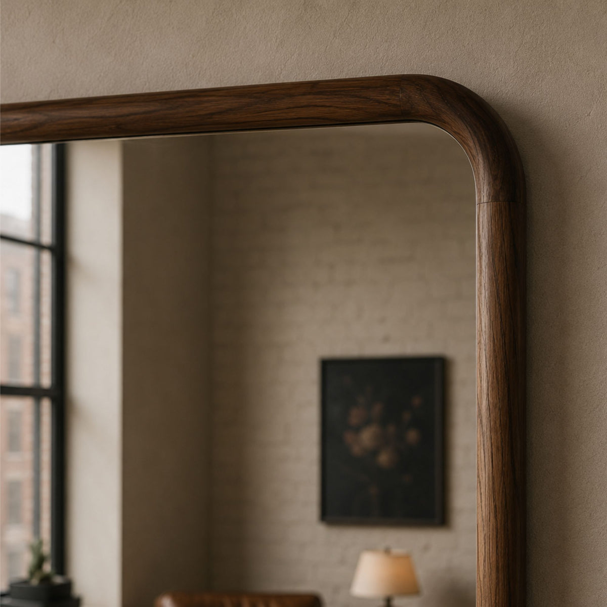 A close-up of a large wooden mirror with a rounded top corner and a dark brown wood grain frame, mounted on a textured beige wall. The reflection shows a softly blurred interior room with a window, brick wall, and lamp.