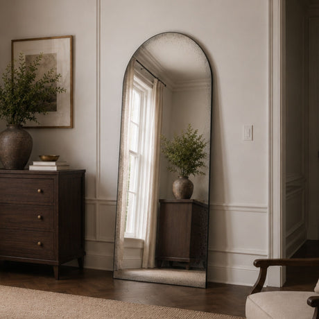 An arched floor mirror with a distressed silver frame stands in a styled room corner, reflecting a window and plant. The mirror is positioned next to a dark wood dresser and framed wall art, with a neutral beige wall and wood floor in the background.