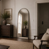 An arched full-length floor mirror with a distressed silver frame, leaning against a white paneled wall in a styled living room.