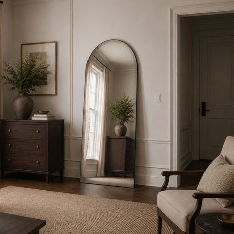 An arched full-length floor mirror with a distressed silver frame, leaning against a white paneled wall in a styled living room.