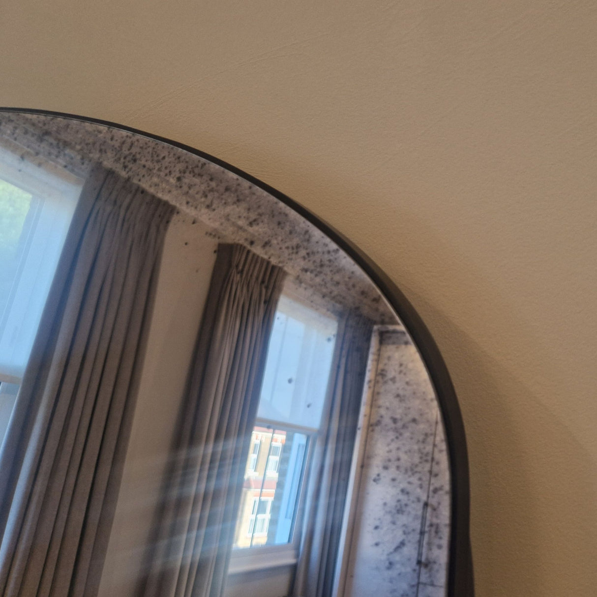 An arched wall mirror with a dark frame reflecting gray curtains and a window, mounted on a textured beige wall.