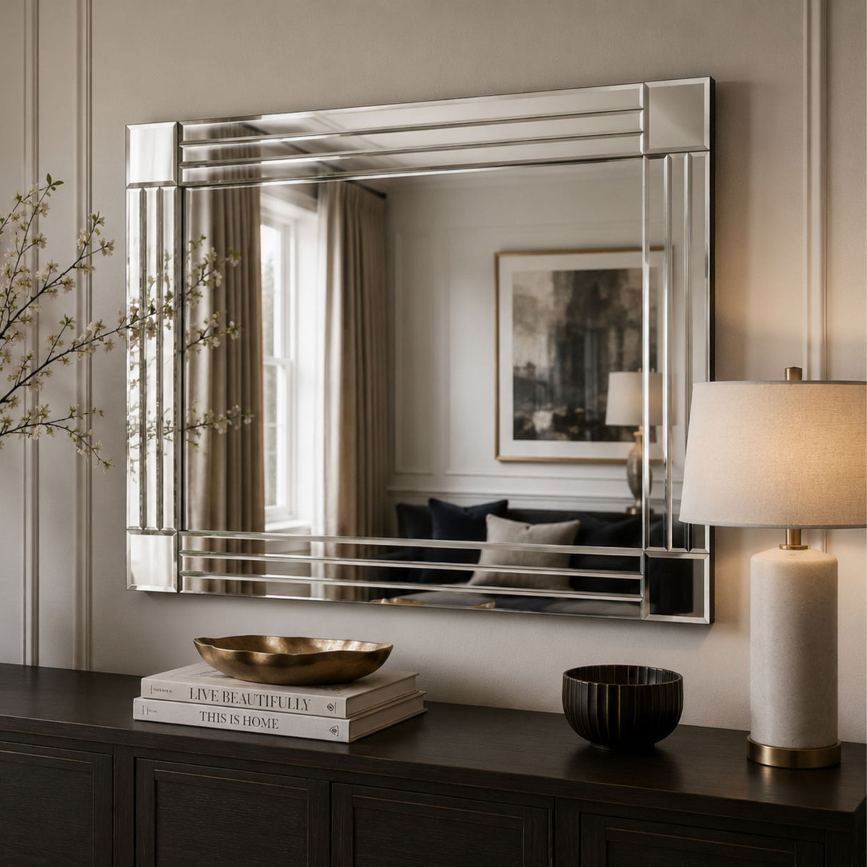 A large rectangular wall mirror with a polished silver frame and horizontal bar accents, mounted on a beige paneled wall above a dark wood console table. The mirror reflects a living room interior, and the scene is styled with a table lamp, books, and decorative bowls.