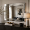 A large rectangular wall mirror with a polished silver frame and horizontal bar accents, mounted on a beige paneled wall above a dark wood console table. The mirror reflects a living room interior, and the scene is styled with a table lamp, books, and decorative bowls.