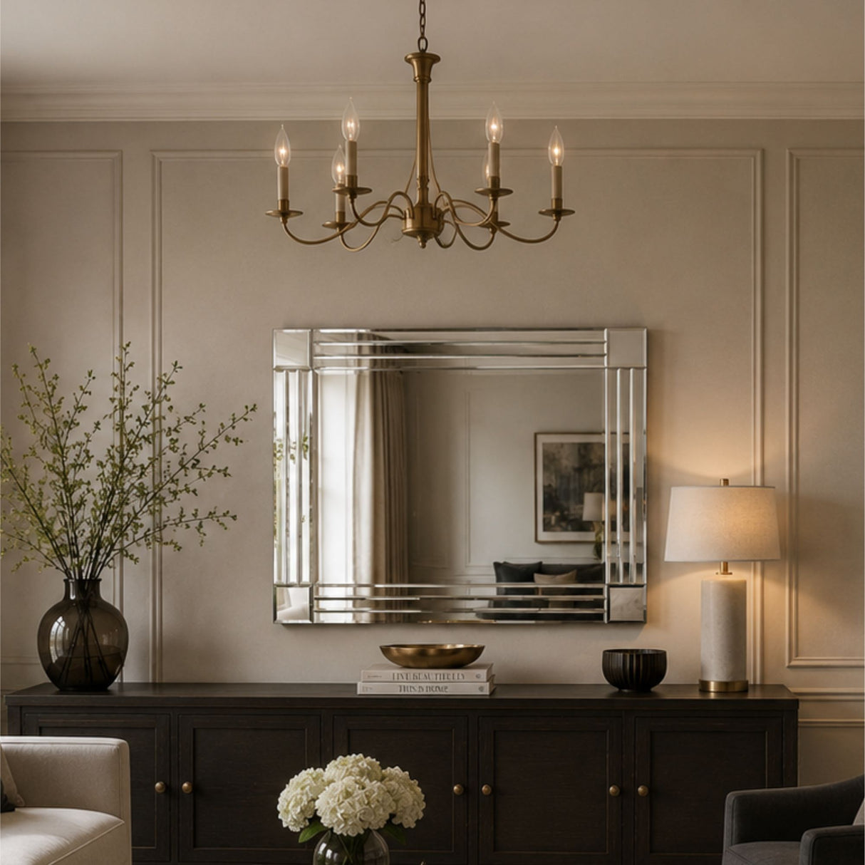 A large rectangular wall mirror with a polished silver frame hangs above a dark wood console table in a styled interior. The room has beige paneled walls, a brass chandelier, a lit table lamp, a vase with branches, and floral arrangements.