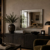 A rectangular wall mirror with a silver-toned beveled frame hangs above a dark wood console in a styled interior. The room includes a large vase with greenery, books, a black bowl, a lit table lamp, and part of an upholstered chair.