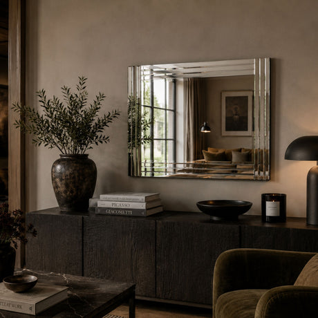 A rectangular wall mirror with a silver-toned beveled frame hangs above a dark wood console in a styled interior. The room includes a large vase with greenery, books, a black bowl, a lit table lamp, and part of an upholstered chair.