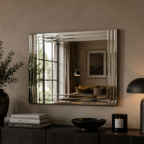 A rectangular wall mirror with a polished silver frame hangs on a warm beige wall above a dark console table. The room setting includes a plant, books, a bowl, and a lit lamp, with the mirror reflecting a cozy interior scene.
