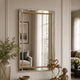 A rectangular wall mirror with a slim gold inner frame and a silver outer border, mounted on a beige wall in a styled interior setting with decorative objects and a reflection of a room behind it.
