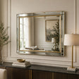 A rectangular wall mirror with a gold-toned frame and mirrored glass, styled in a classic interior setting above a dark wood console table with a lamp, books, and a vase.