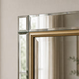 Close-up of a rectangular wall mirror with a polished silver-toned frame and a gold inner border, reflecting a softly lit interior.