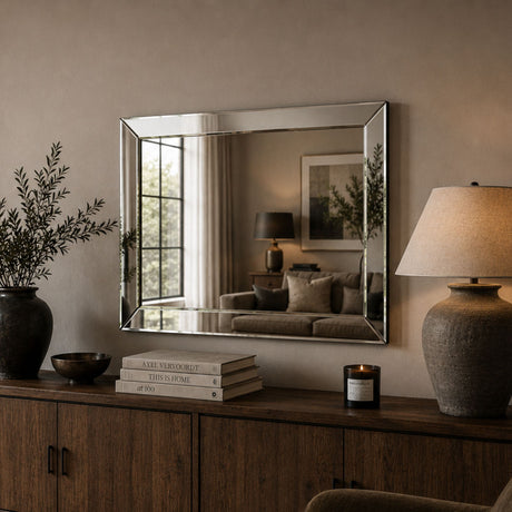 A rectangular wall mirror with a thin silver frame hangs above a wooden console table in a styled interior. The mirror reflects a softly lit living room, while the surrounding scene includes a vase with branches, books, a candle, and a table lamp.