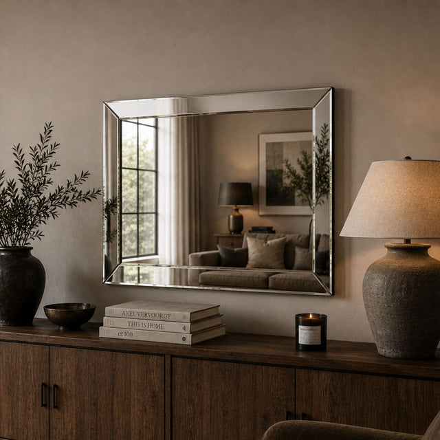 A rectangular wall mirror with a thin silver frame hangs above a wooden console table in a styled interior. The mirror reflects a softly lit living room, while the surrounding scene includes a vase with branches, books, a candle, and a table lamp.