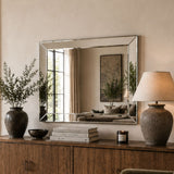 A large rectangular wall mirror with a silver-toned frame hangs above a wooden cabinet in a styled interior. The mirror reflects a living room scene, while the room itself is decorated with a vase of greenery, books, a candle, and a table lamp.