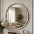 A large round wall mirror with a ribbed gold-toned frame hangs on a light beige paneled wall. The mirror reflects a softly lit living room with a sofa, curtains, and a plant.