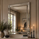 A large rectangular wall mirror with a polished silver frame hangs on a beige paneled wall above a mantel. The mirror reflects a softly lit interior with curtains, a lamp, and framed art, while the surrounding scene includes a potted plant, a bowl, and two lit candles.